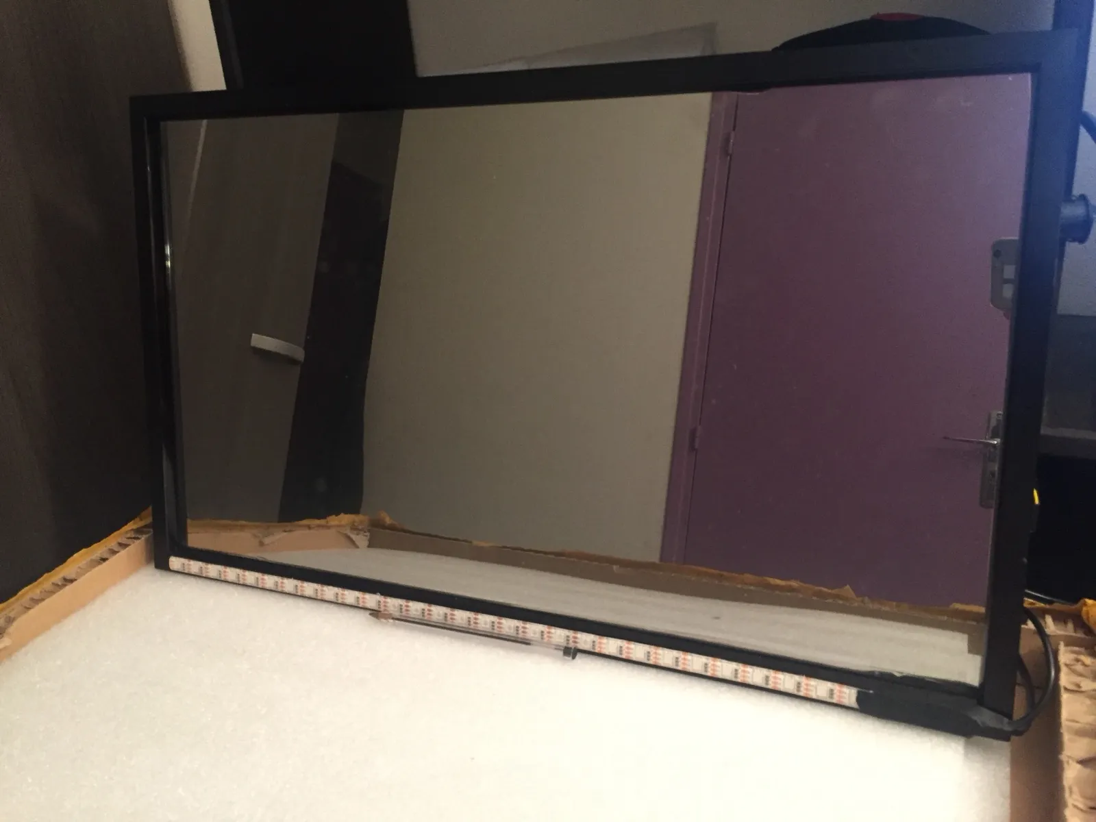 Glace sans tain montée dans un cadre d’aluminium noir, posée à la verticale sur un bloc de mousse blanche ; un ruban de LED adhésif court le long du bord inférieur intérieur.